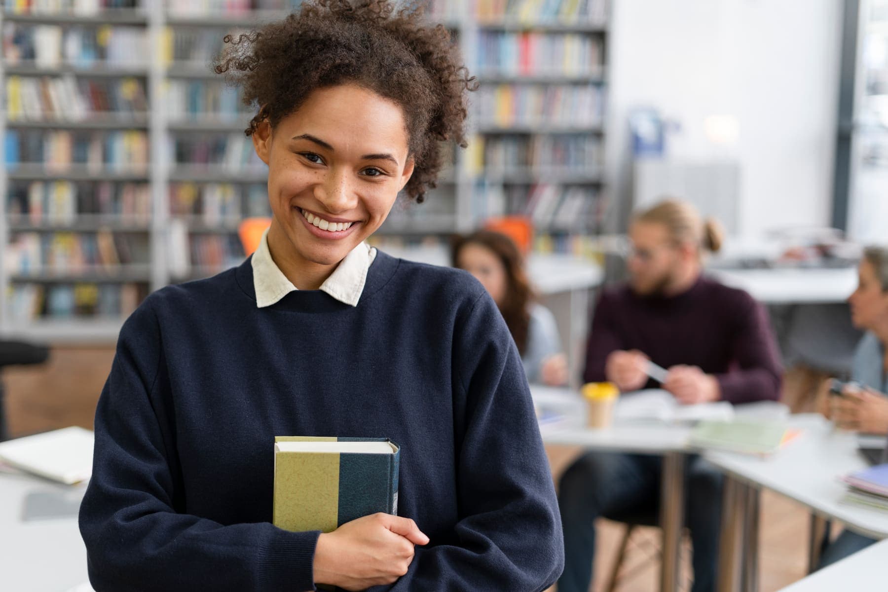 A student in a navy school jumper and collared shirt holding a book in her school library, with classmates studying behind her